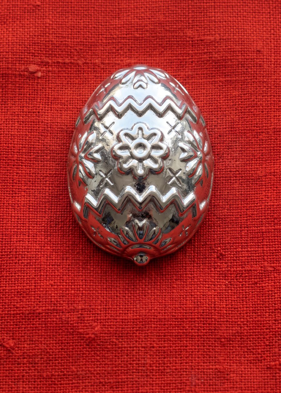 a chrome egg standing upright, covered in stitched folk rosettes and zigzag bands